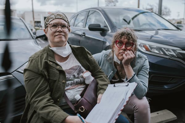 Tilly and Susan sitting down in a parking lot, looking up at the camera monitor during filming on “Long Away”